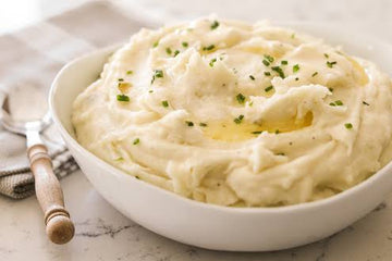 A white ceramic bowl filled with creamy mashed potatoes topped with melted butter and chopped chives, set on a light marble surface with a spoon and napkin nearby.