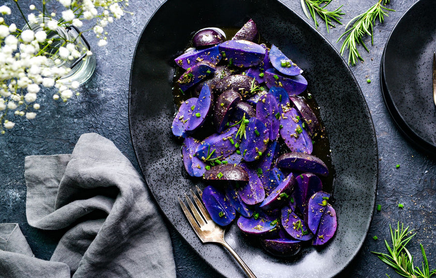 Purple potatoes sliced and cooked with oil, herbs, and lemon zest, served on a dark oval platter with a fork, set on a dark textured tabletop with rosemary, baby's breath, and a gray napkin.