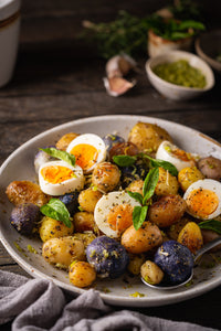 A close-up of a plated cooked potato salad on a rustic dark wooden table. The dish contains mixed baby and medium potatoes, halved hard-boiled eggs, fresh green basil leaves, and olive oil dressing, served on a light gray ceramic plate with a spoon.