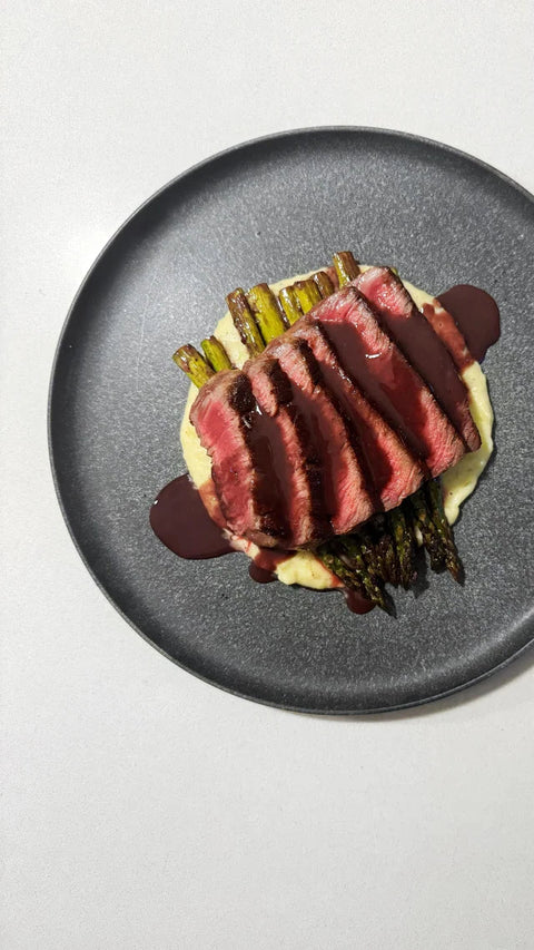 A plated gourmet dish featuring sliced raw beef with a dark red sauce, topped with green asparagus and served over blue moon mashed potatoes on a dark gray plate against a white background.